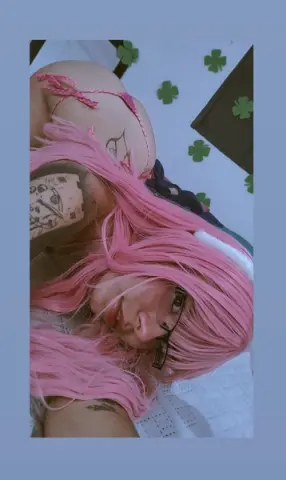 Woman with pink hair and tattoos wearing pink bikini in a room with clover decorations.