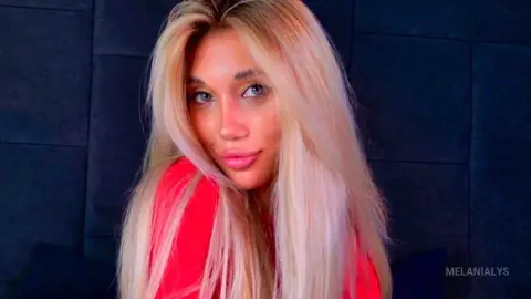 Blonde woman in a red top smiling against a dark textured wall background.