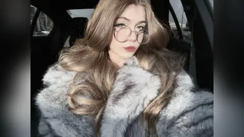 Young woman with long blonde hair wearing a grey faux fur coat and round glasses inside a car.