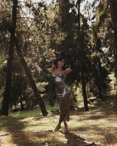 Elegant woman in a shimmering silver satin dress posing in a sunlit, moody forest setting.