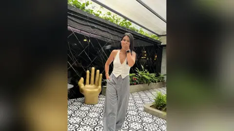 Stylish woman in a white sleeveless top and grey trousers posing by a gold hand sculpture in a modern patio.