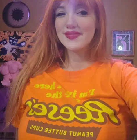 Smiling redhead woman in an orange graphic t-shirt posing in a cozy room with artistic wall decor.