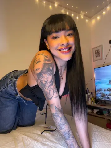 asialovee smiles while kneeling on a bed, showing off her large arm tattoos and black crop top.