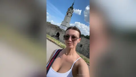 Young woman in a white tank top and sunglasses smiling in front of a historic European church tower.