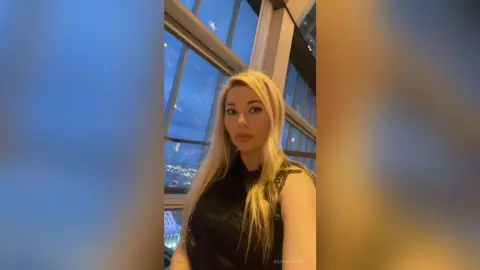 Blonde woman in a black sleeveless top posing by a high-rise window overlooking a city skyline at twilight.
