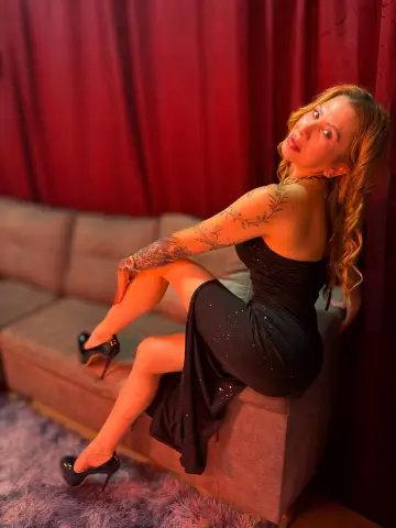 Elegant woman in a black evening gown sitting on a sofa against red curtains in a moody setting.