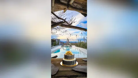 Elegant two-tier black and gold cake on a wooden table by a luxury poolside villa under a blue sky.