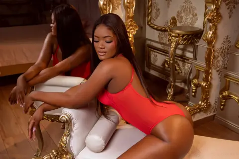 Beautiful woman in a red bodysuit relaxing on a white vintage chair in a luxurious gold-accented room.