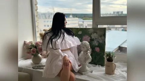 TessaWinsent — TessaWinsent sits by a window in a white ruffled dress with her long black hair flowing down her back.