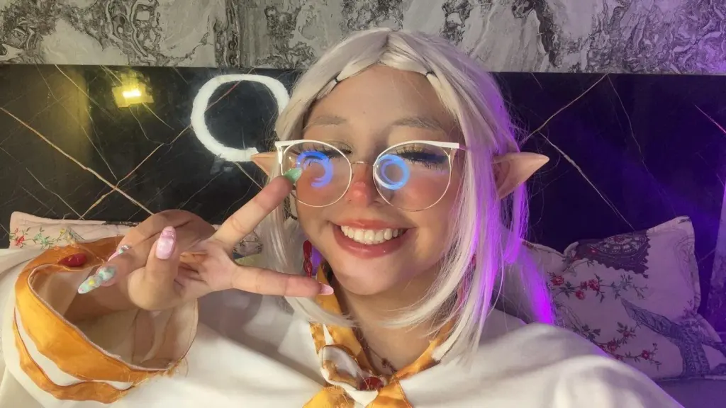 EmyGom3zz poses with long blonde hair and pink-tinted glasses, wearing a red top that shows her cleavage.
