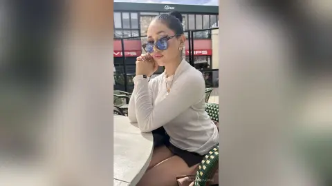 NashiroSakurai sits at a round table wearing a white long-sleeved top and sunglasses, resting her chin on her hands.
