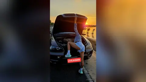 Young woman in denim shorts and grey shirt posing by a car with an open hood during a golden sunset by a lake.