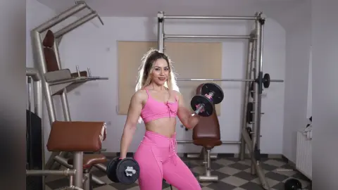 Fit blonde woman in pink gym set lifting dumbbells in a modern bright fitness studio.