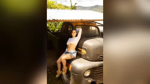 JuliahLewis leans against a vintage black truck wearing a white long-sleeve crop top and denim shorts.