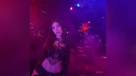 KateGale poses in a dark, smoky setting wearing a black lace crop top and dark pants, lit by red and blue neon lights.