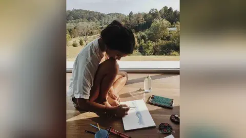 Young woman in a white shirt painting on a sketchbook while sitting on a wooden floor by a scenic forest window.