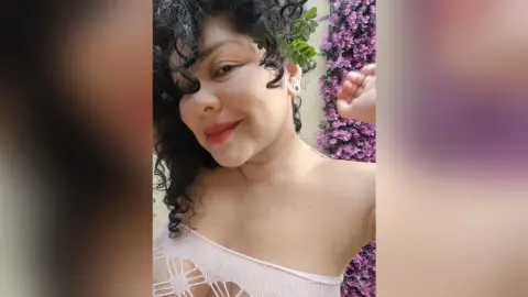 Smiling woman with curly hair wearing a white lace top in a garden setting with pink flowers.