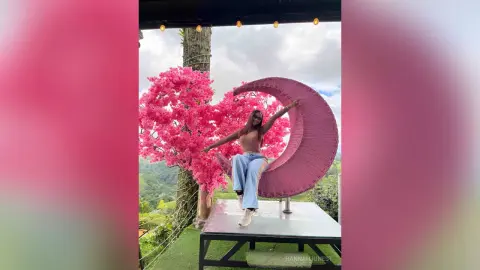 Stylish woman in light blue jeans sitting on a crescent moon swing near pink cherry blossom trees outdoors.