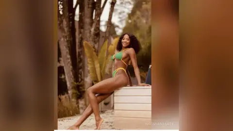 Smiling Black woman in a green bikini posing outdoors in a tropical garden setting.