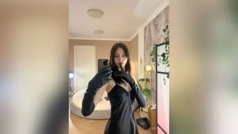 Young woman in a sleek black leather dress and gloves taking a moody mirror selfie in a modern bedroom.