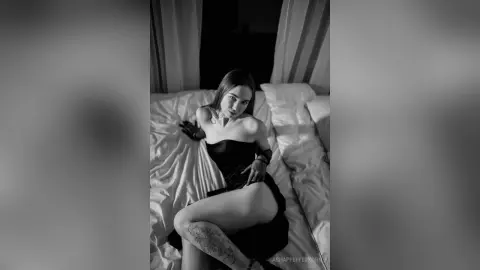 Moody black and white portrait of a tattooed woman in a black strapless dress relaxing on a white bed.