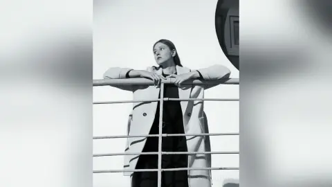 Elegant woman in a long coat leaning on a balcony railing against a bright sky in a moody black and white portrait.