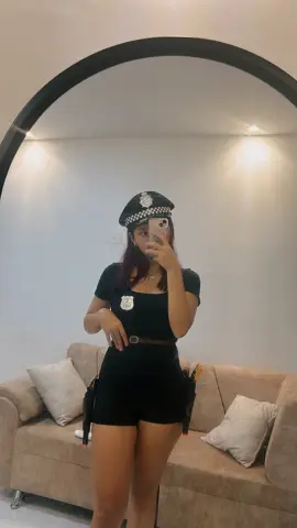 AlyMartini poses in a black police officer costume and hat, standing in front of a large mirror in a living room.