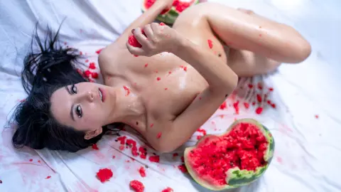 Nude woman lying on white fabric surrounded by red watermelon pieces in a bright, summery setting.