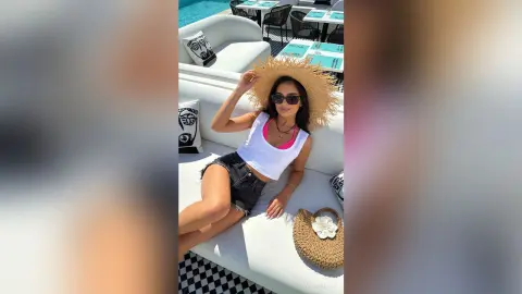 Stylish woman in white tank top and denim shorts relaxing on a white lounge chair by a sunny poolside.