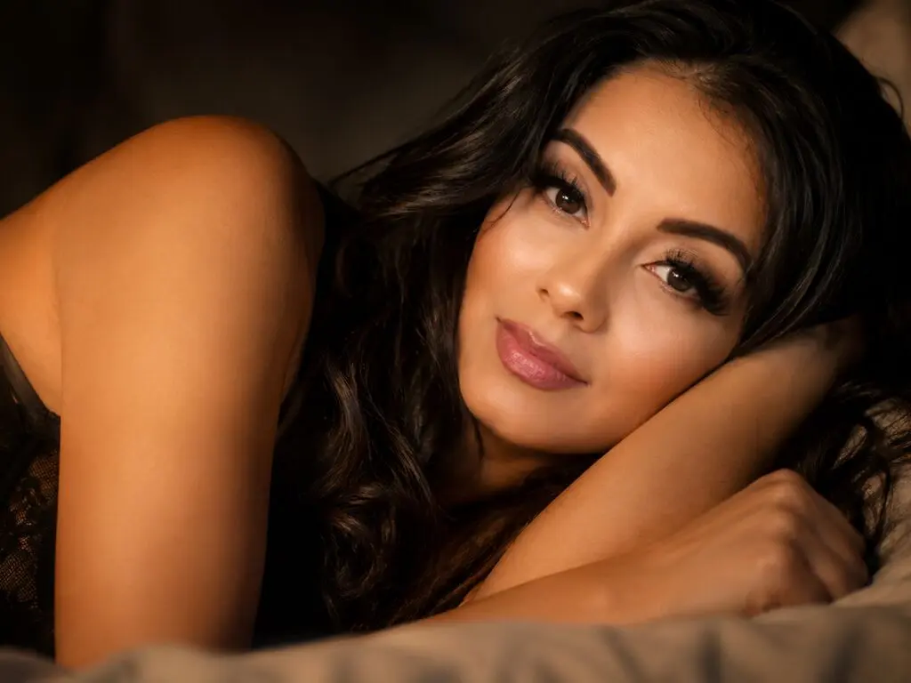AlessaWaldorf lies on a bed with long dark hair and a soft smile, looking directly at the camera.