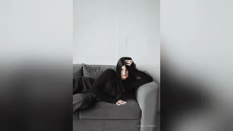 Moody portrait of a woman in a black long-sleeve top relaxing on a grey sofa in a minimalist room.