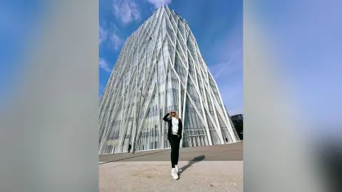 MilenaBia stands in front of a large, modern glass building, wearing a white tank top, black pants, and white sneakers.