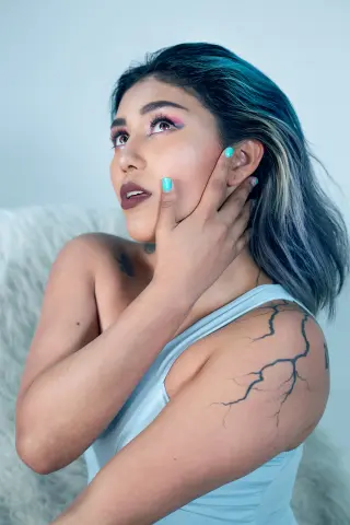 Dreamy portrait of a woman with blue hair and tattoos wearing a light blue tank top against a white background.