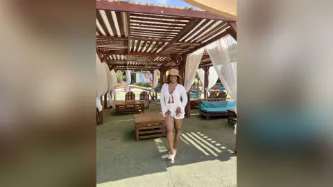 Smiling woman in a white linen outfit and sun hat relaxing under a wooden pergola at a sunny outdoor resort.