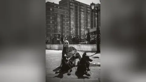 Stylish woman in a sequined outfit sitting with two black dogs in an urban city setting, black and white mood.