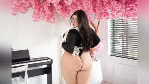 Curvy woman in a black maid outfit posing in a bright room with pink cherry blossom decor.