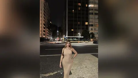 Elegant woman in a beige bodycon dress standing on a city sidewalk at night near urban buildings.