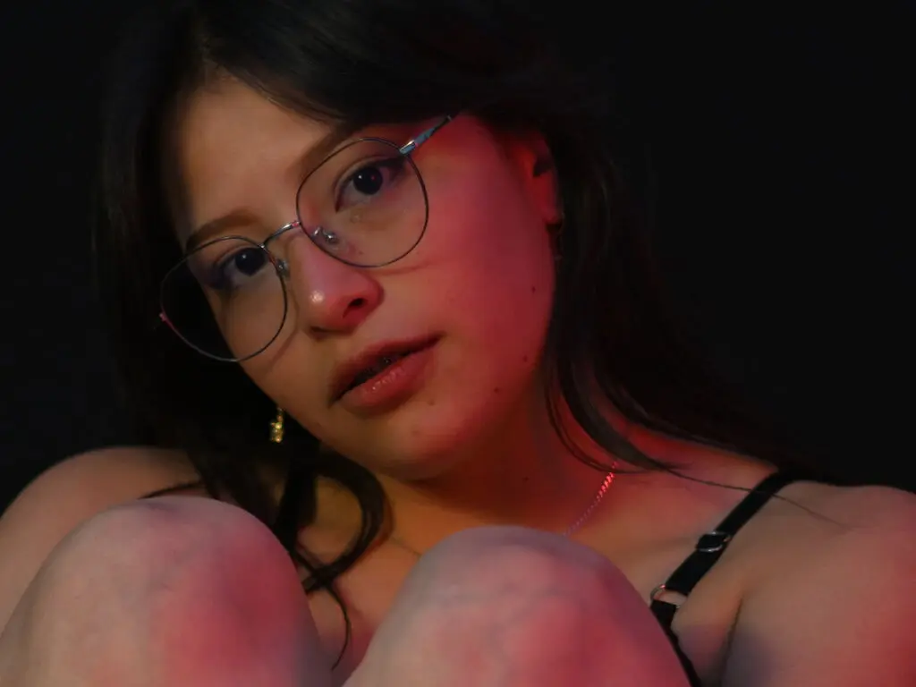 Young woman with glasses and dark hair in moody red lighting wearing a black strap top.