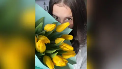 Young woman with long dark hair peeking through a vibrant bouquet of bright yellow tulips in a soft indoor setting.