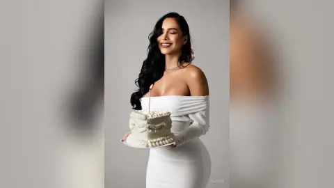 Alison smiles while holding a white frosted cake with a single lit candle, wearing a white off-the-shoulder dress.