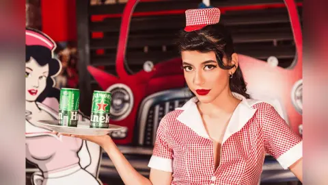 JessieLiu wears a red and white checkered waitress outfit while holding a tray with two green cans in front of a red truck.