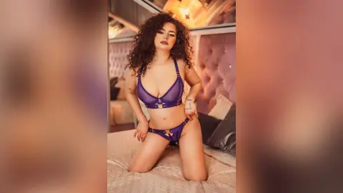 Curvy woman in purple lace lingerie posing on a bed in a cozy, dimly lit bedroom.