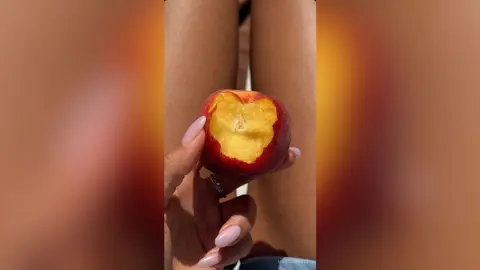MarisaGrace holds a sliced red peach in front of her legs, showing the yellow fruit inside.