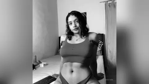 Young woman in a ribbed crop top sitting on a bed in a moody black and white indoor portrait.