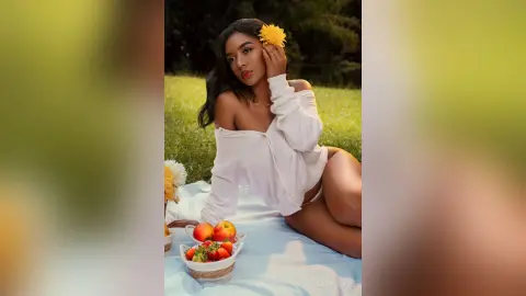 Beautiful woman in a white off-the-shoulder shirt relaxing on a picnic blanket with fruit in a sunny garden.