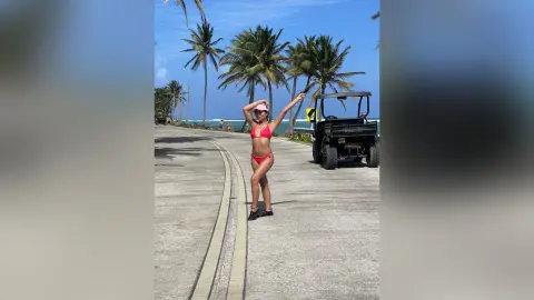 Young woman in a red bikini posing on a sunny tropical road next to a palm tree and off-road vehicle.