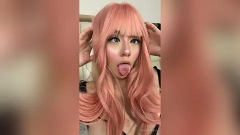Playful girl with long pink hair and bangs sticking her tongue out in a casual indoor setting.