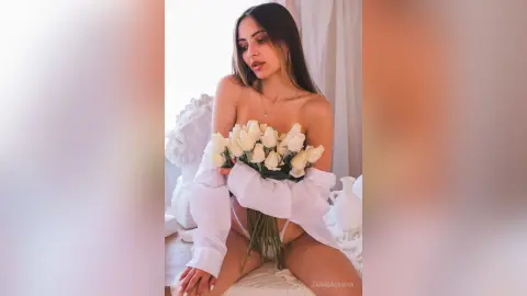 Young woman in a white blouse holding a large bouquet of white roses in a bright, airy room.