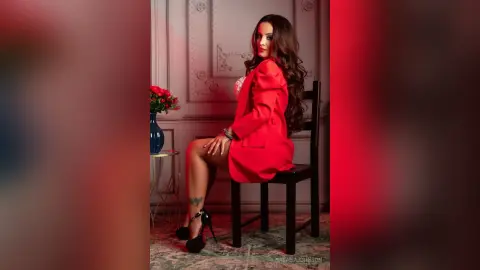 Elegant woman in a red blazer and black heels sitting on a chair in a classic room with a moody atmosphere.