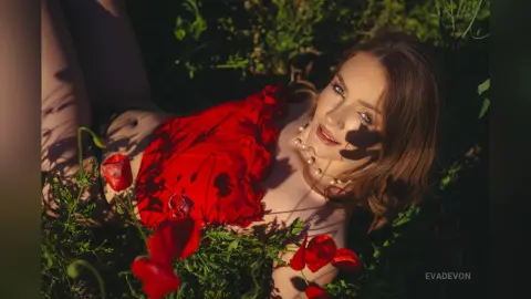 Young woman in a red ruffled top lying in lush green foliage under warm sunlight for a romantic summer portrait.